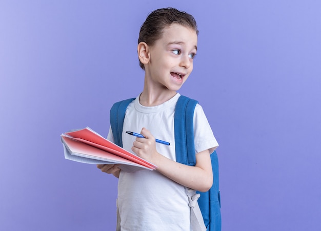 Impressed little boy wearing backpack standing in profile view holding pen and note pad looking at side isolated on purple wall with copy space