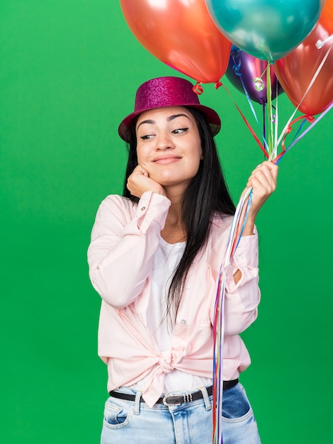 Colpito guardando lato giovane bella ragazza che indossa un cappello da festa tenendo palloncini mettendo la mano sulla guancia isolata sul muro verde