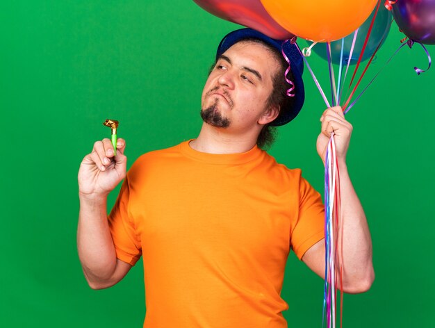 Impressed looking side young man wearing party hat holding balloons with party whistle isolated on green wall