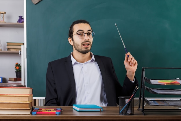 Impressed male teacher wearing glasses points with pointer stick at blackboard sitting at table with school tools in classroom