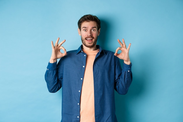 Impressed man praising something awesome, showing okay signs and looking amazed, complimenting good thing, standing on blue background.