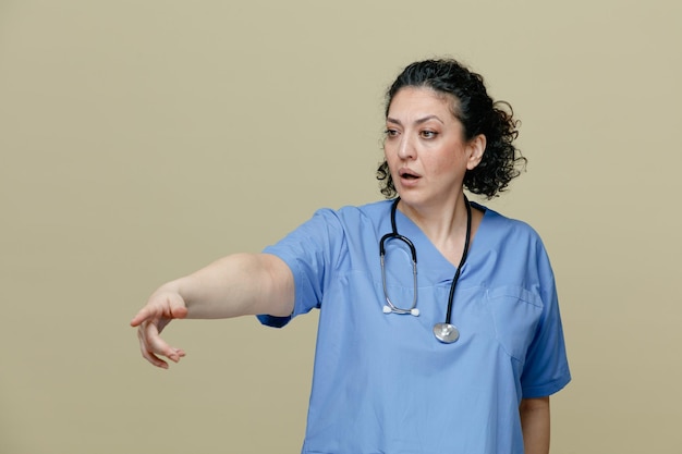 Impressed middleaged female doctor wearing uniform and stethoscope around neck looking and pointing at side isolated on olive background