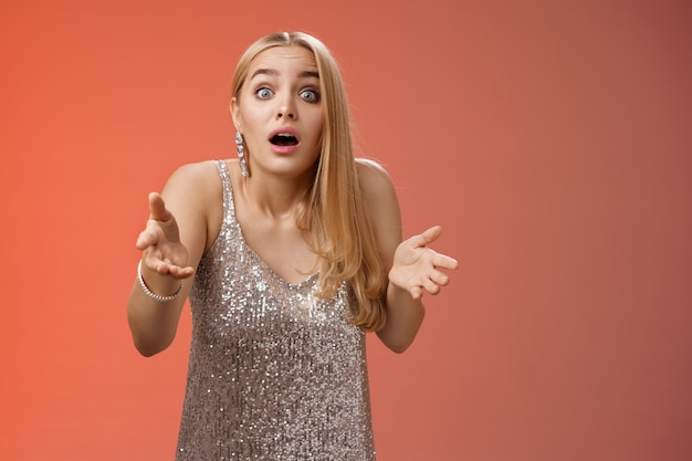 Impressed overwhelmed blond woman retelling exciting story gesturing astonished widen eyes shrugging explaining what happened talking passionately in silver glamour dress standing red background.