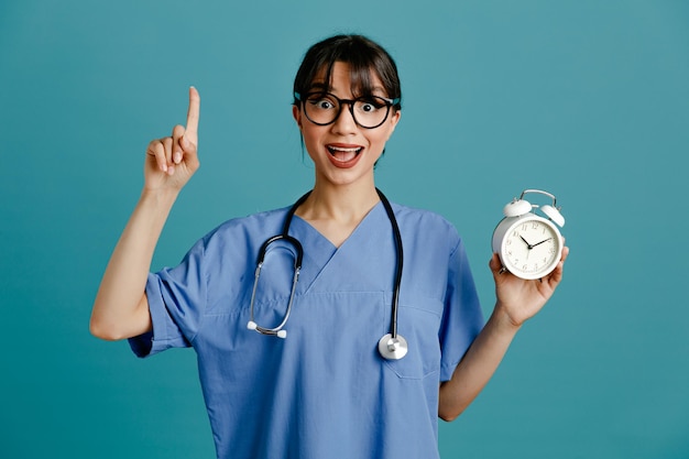 impressed points at up holding alarm clock young female doctor wearing uniform fith stethoscope isolated on blue background