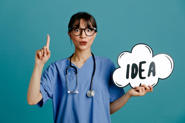 Impressed points at up holding idea bubble young female doctor wearing uniform fith stethoscope isolated on blue background