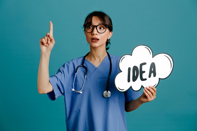 Impressed points at up holding idea bubble young female doctor wearing uniform fith stethoscope isolated on blue background