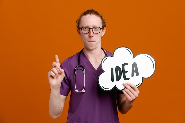 Impressed points at up holding idea bubble young male doctor wearing uniform with stethoscope isolated on orange background