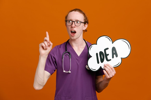 Impressed points at up holding idea bubble young male doctor wearing uniform with stethoscope isolated on orange background