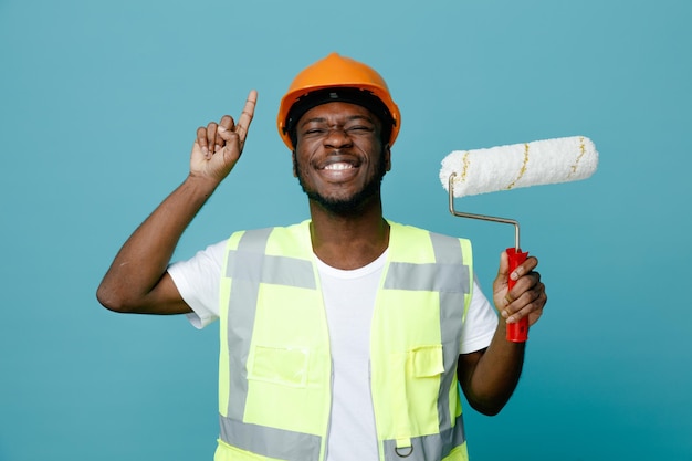 Impressed points at up young african american builder in uniform holding roller brush isolated on blue background