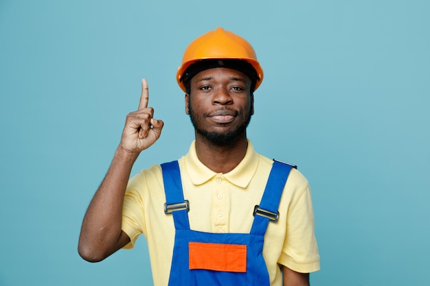 Impressed points at up young african american builder in uniform isolated on blue background