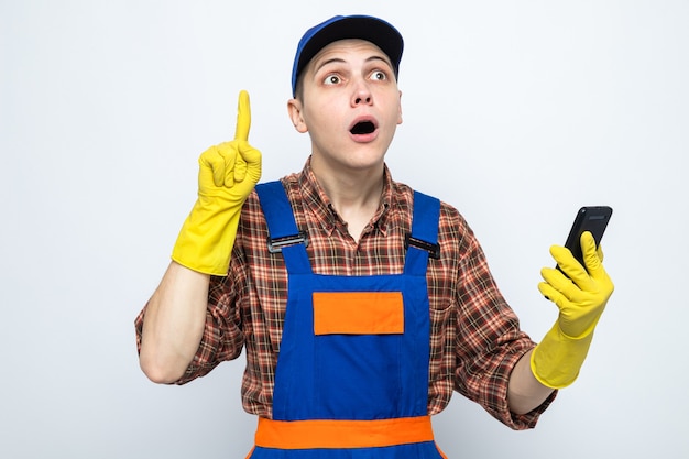 Impressed points at up young cleaning guy wearing uniform and cap with gloves holding phone 