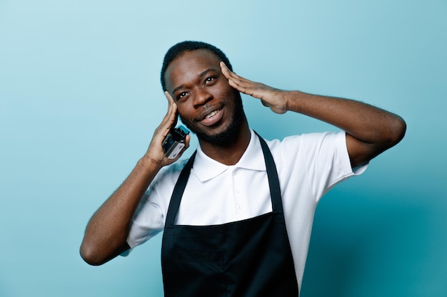 Impressed putting hands on head holding hair clippers young african american barber in uniform isolated on blue background