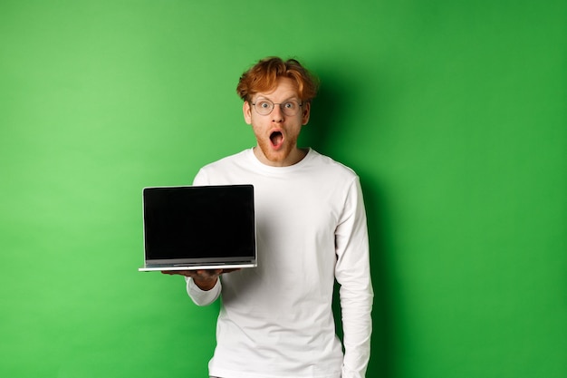 안경을 쓴 빨간 머리 남자는 온라인 프로모션을 본 후 턱을 떨어뜨리고 노트북 화면을 보여주고 믿을 수 없다는 표정으로 카메라를 응시하고 녹색 배경 위에 서 있습니다.
