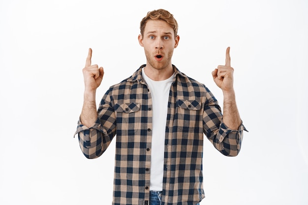 Impressed redhead man drop jaw, gasping in awe and pointing fingers up at something cool, showing new promo store deal, best offer, standing over white wall.