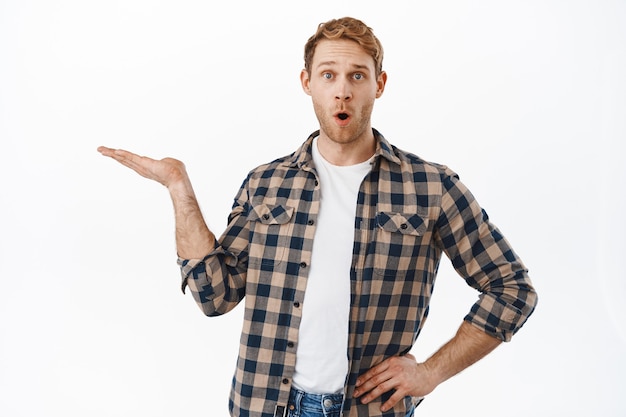 Impressed redhead man say wow while holding an item, raising hand with opened palm, showing an object, product in his arms, standing over white wall