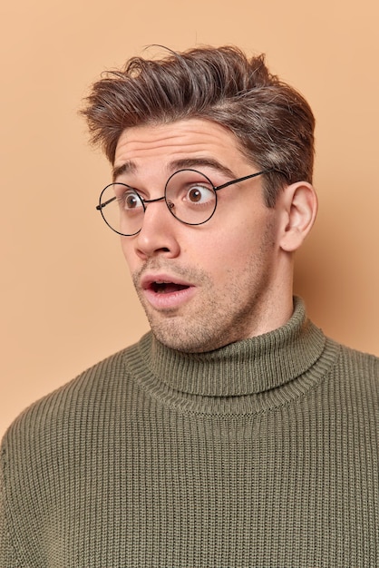 Impressed shocked adult man with dark hair stares stunned away notices horrible thing feels amazed wears spectacles and sweater reacts on amazing news isolated over beige background. Omg concept