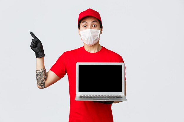 Impressed, shocked asian delivery guy in red uniform, cap, holding laptop, showing screen advertisement, pointing upper left corner at online shopping site, courier deliver clients orders