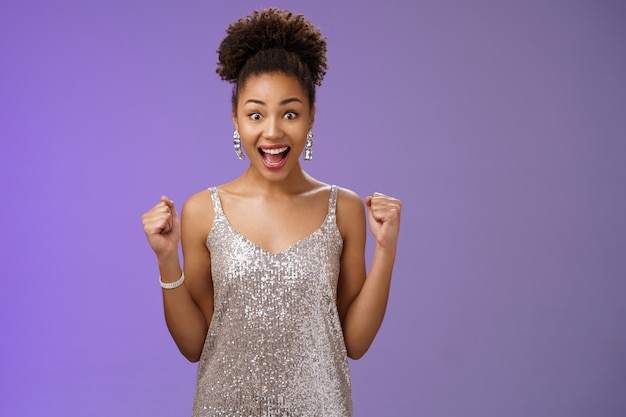 Impressed surprised thrilled joyful charming elegant african-american woman winning money casino achieve success yelling happily raise fists victory triumph gesture cheering gladly blue background.