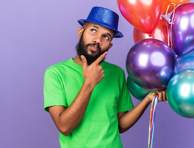 Impressed young afro-american guy wearing party hat holding balloons putting hand on chin isolated on blue wall