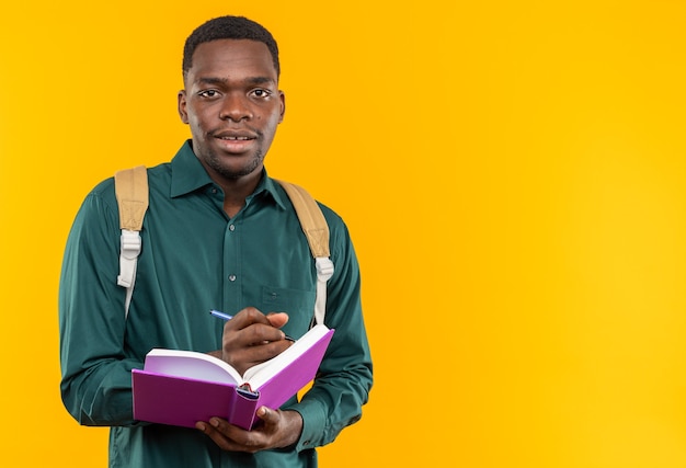 Впечатленный молодой афро-американский студент с рюкзаком, держащим книгу и ручку, изолированные на оранжевой стене с копией пространства