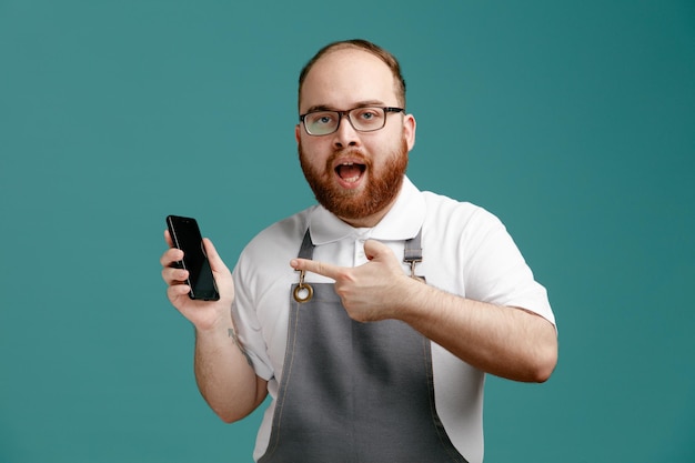 Impressed young barber wearing uniform and glasses holding and pointing at mobile phone looking at camera isolated on blue background