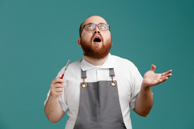 Impressed young barber wearing uniform and glasses holding straight razor looking up showing empty hand isolated on blue background