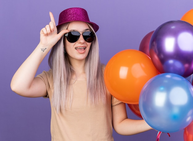 Impressed young beautiful girl wearing dental braces and party hat with glasses holding balloons with gift bag points up isolated on blue wall