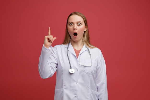 Impressed young blonde female doctor wearing medical robe and stethoscope around neck pointing up 