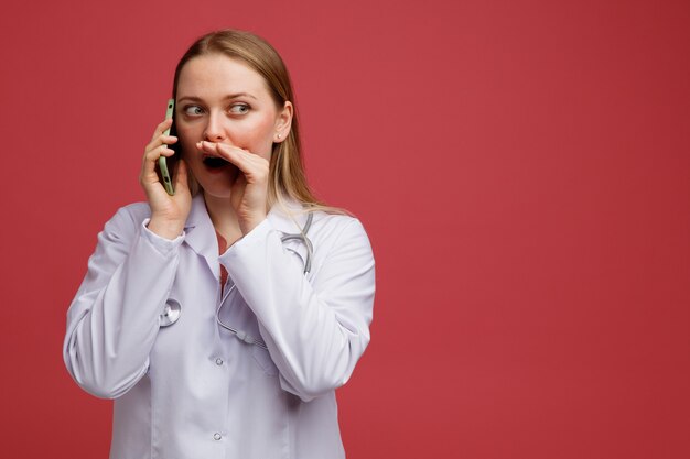 Impressed young blonde female doctor wearing medical robe and stethoscope around neck whispering on phone looking at side 