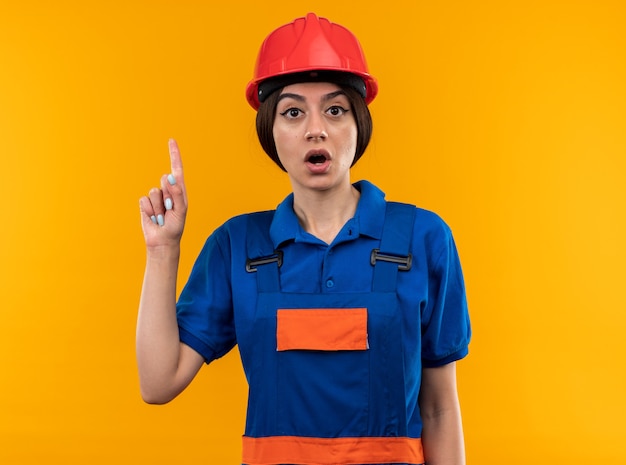 Impressed young builder woman in uniform points at up 