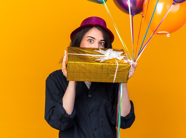 Impressed young caucasian party girl wearing party hat holding bunch of balloons and gift package from behind package isolated on orange wall