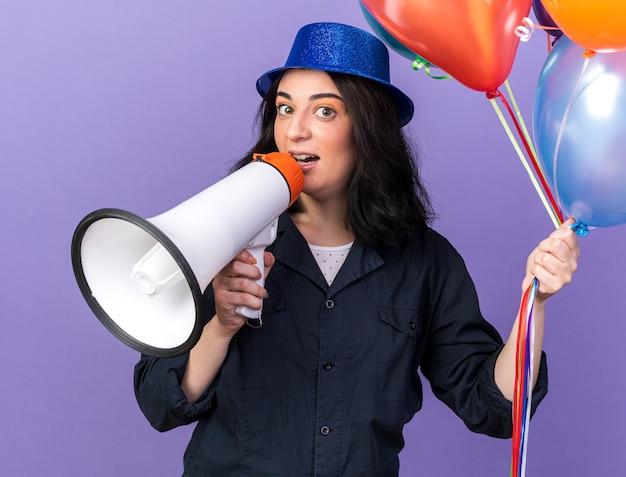 Impressed young caucasian party girl wearing party hat holding bunch of balloons  talking by speaker isolated on purple wall