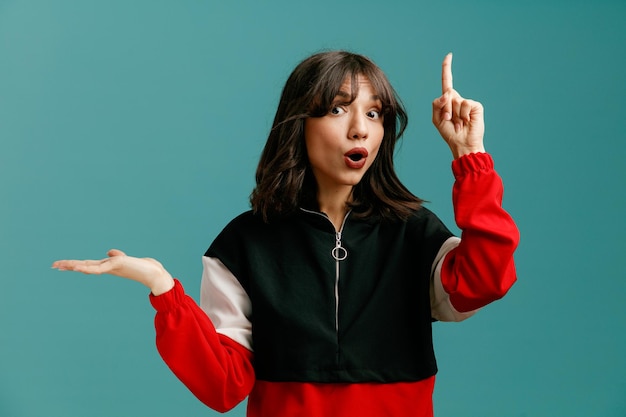 Impressed young caucasian woman looking at camera showing empty hand pointing up isolated on blue background