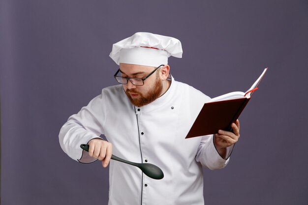 Impressed young chef wearing glasses uniform and cap holding note pad and solid spoon looking down isolated on purple background