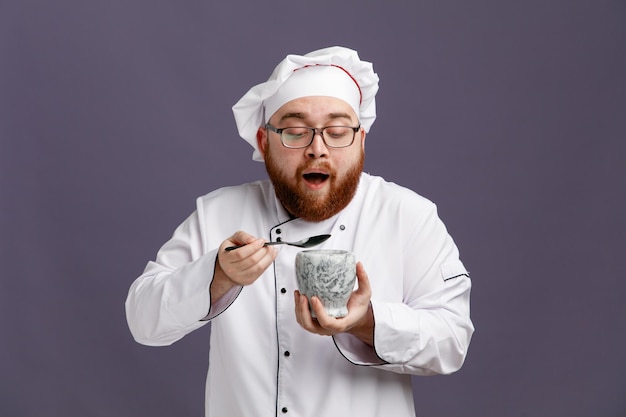 Impressed young chef wearing glasses uniform and cap holding spoon and bowl looking at bowl isolated on purple background