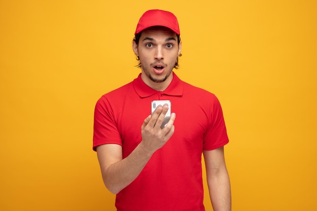 impressed young delivery man wearing uniform and cap holding mobile phone looking at camera isolated on yellow background