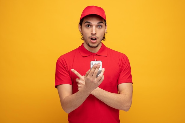 impressed young delivery man wearing uniform and cap holding mobile phone looking at camera pointing to side isolated on yellow background
