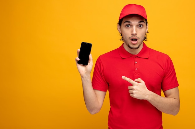 Impressionato giovane fattorino che indossa uniforme e cappuccio guardando la fotocamera che mostra il telefono cellulare che punta su di esso isolato su sfondo giallo con spazio per la copia