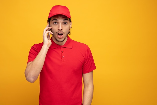 Impressionato giovane fattorino che indossa uniforme e cappuccio guardando la fotocamera parlando al telefono isolato su sfondo giallo