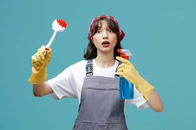 Впечатленная молодая женщина-уборщица в униформе бандана и резиновых перчатках, держащая щетку и моющее средство, смотрящая в камеру, изолированную на синем фоне