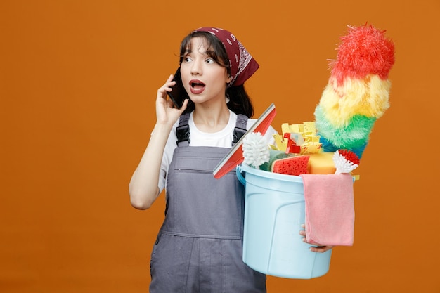 Foto impressionata giovane donna addetta alle pulizie che indossa uniforme e bandana che tiene secchio di strumenti per la pulizia parlando al telefono guardando il lato isolato su sfondo arancione