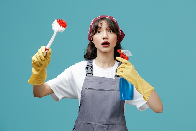 Impressed young female cleaner wearing uniform bandana and rubber gloves holding brush and cleanser looking at camera isolated on blue background