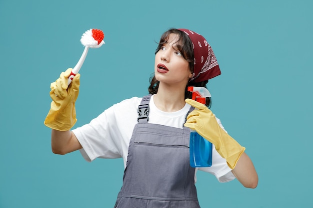 Impressed young female cleaner wearing uniform bandana and rubber gloves holding brush and cleanser looking at side isolated on blue background