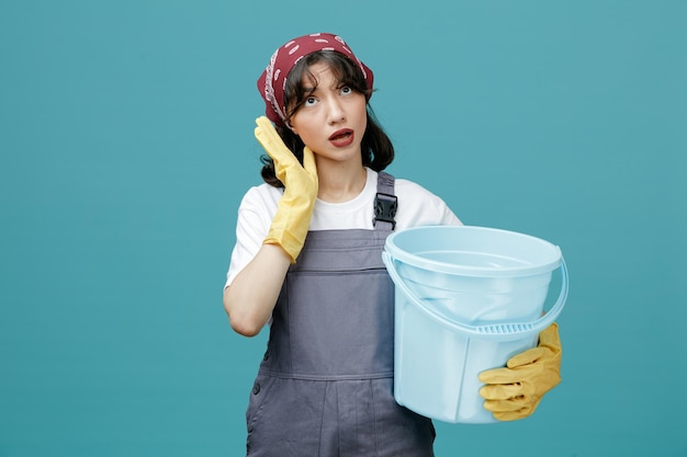 Impressed young female cleaner wearing uniform bandana and rubber gloves holding bucket keeping hand near face looking up isolated on blue background