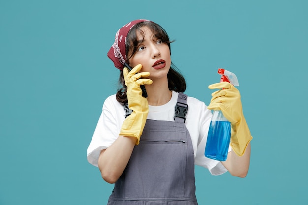 Impressed young female cleaner wearing uniform bandana and rubber gloves holding cleanser looking at side talking on phone isolated on blue background