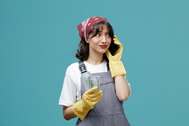 Impressed young female cleaner wearing uniform bandana and rubber gloves holding money looking at side talking on phone isolated on blue background