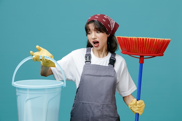 impressed young female cleaner wearing uniform bandana and rubber gloves holding squeegee mop and bucket looking at bucket isolated on blue background