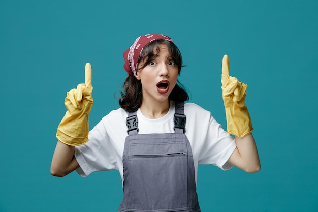 Impressionato giovane donna addetta alle pulizie che indossa bandana uniforme e guanti di gomma guardando la fotocamera che punta le dita verso l'alto isolato su sfondo blu