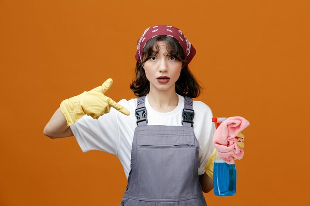 Impressed young female cleaner wearing uniform rubber gloves and bandana holding cleanser and cloth duster pointing at them looking at camera isolated on orange background