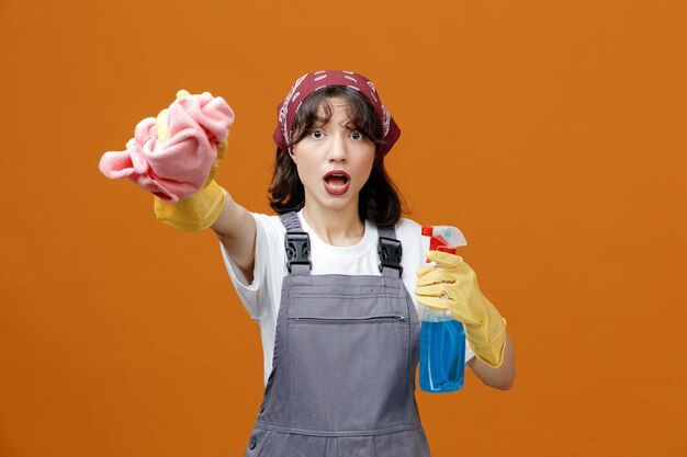 Photo impressed young female cleaner wearing uniform rubber gloves and bandana holding cleanser stretching cloth duster out towards camera looking at camera isolated on orange background